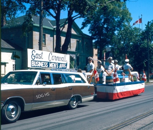 A parade float in East Cornwall features a car towing a platform with people in uniforms holding instruments. A sign reads "East Cornwall Business Men's Assoc."