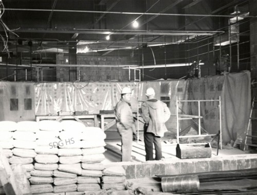 Two construction workers in hard hats stand in a spacious, indoor construction site. They are surrounded by scaffolding and stacked cement bags. The area is dimly lit, creating a focused and industrious atmosphere.