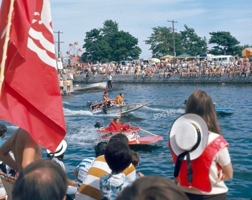 Boat race on a sunny day with two speedboats on water, surrounded by a large crowd. People are watching enthusiastically, with flags waving. Energetic atmosphere.