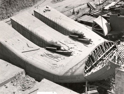 Aerial view of a large, partially constructed concrete dam with exposed rebar and scaffolding. Surrounding construction site with tools and debris.