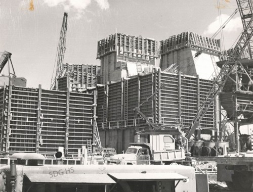 Black-and-white photo of a large construction site with towering scaffolding, cranes, trucks, and workers. The scene conveys industrial progress.