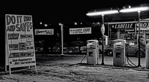 Vintage black-and-white scene of an old gas station at night, featuring retro gas pumps, signage for discounts, and a classic roadside atmosphere.