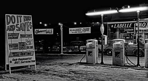 Vintage black-and-white scene of an old gas station at night, featuring retro gas pumps, signage for discounts, and a classic roadside atmosphere.