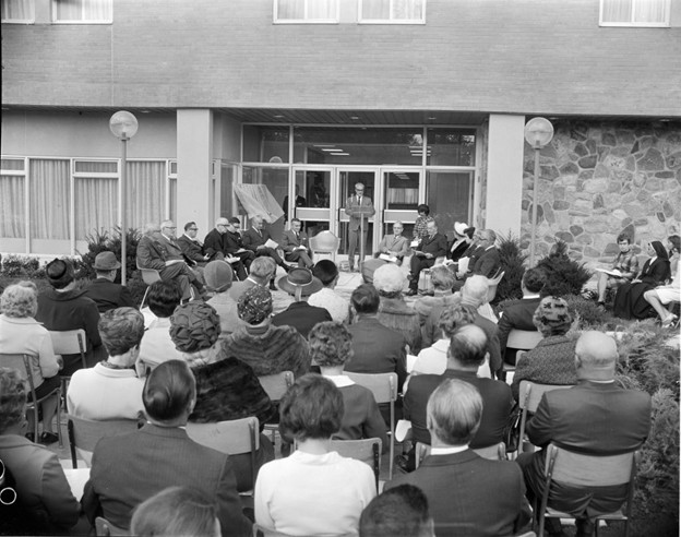 Black and white photo of a formal outdoor gathering, featuring a speaker at a podium in front of an audience. Attendees sit on chairs, dressed in 1950s attire.
