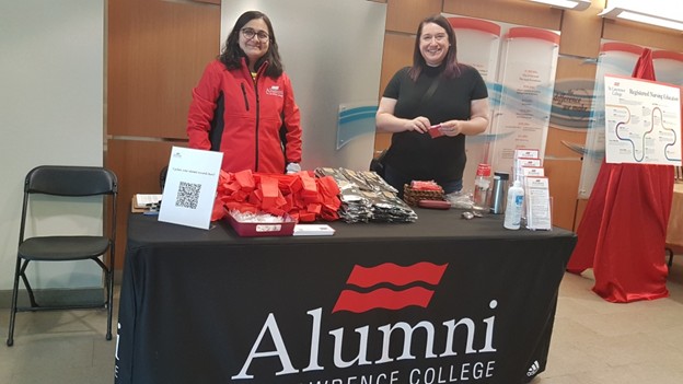Two people stand behind a table with a black tablecloth labeled "Alumni Lawrence College." The table has red gift bags, pamphlets, and a QR code. The atmosphere is welcoming.