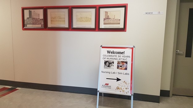 Hallway displaying four framed historical photos and documents in red frames on a wall. A signboard directs to the "Nursing Lab/Sim Labs" for a 50th-anniversary celebration of nursing at SLC.