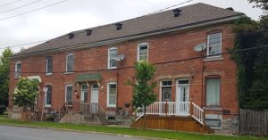 A row of connected brick townhouses with grey roofs and small front porches. Trees and wires line the street, under an overcast sky.