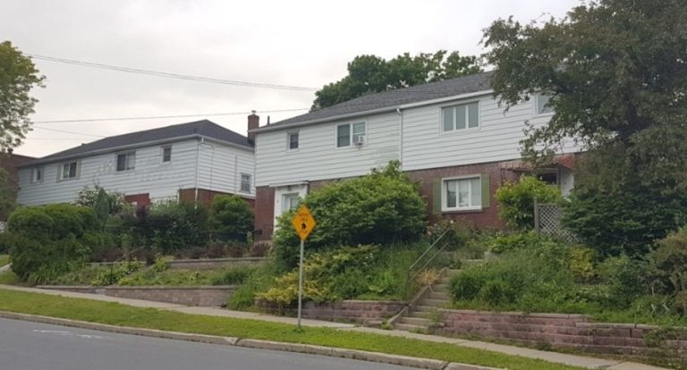 Two side-by-side suburban houses with white siding and red brick accents, surrounded by lush green bushes and a tree. A pedestrian crossing sign is in the foreground.