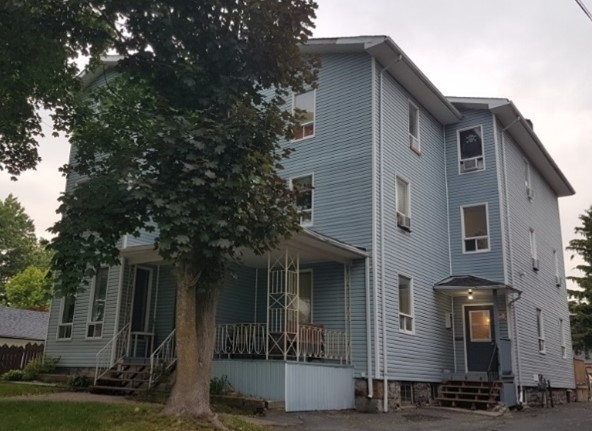 Three-story blue house with white trim, surrounded by trees. Multiple windows and a porch give it a residential feel. Overcast sky suggests a calm, quiet mood.