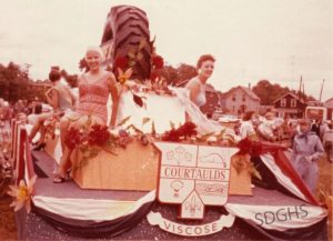 Vintage parade float with a "Courtaulds Viscose" sign, adorned with red flowers. Women in swimsuits smile, evoking a joyful, festive atmosphere.