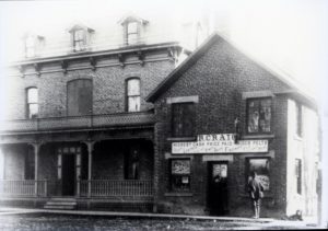 This photo from the Museum’s Daly Collection, appears to show some of the buildings that once occupied the Craig Pitt Street site. The collection contains 108 local photographs dating between 1893 and 1901. Historic black-and-white photo of a brick building with a sign reading "R. Craig." Two men stand outside. The scene has a vintage, early 20th-century feel.
