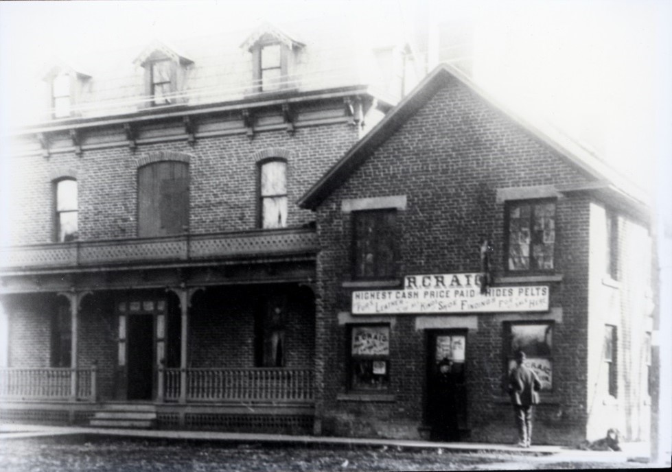 This photo from the Museum’s Daly Collection, appears to show some of the buildings that once occupied the Craig Pitt Street site. The collection contains 108 local photographs dating between 1893 and 1901. Historic black-and-white photo of a brick building with a sign reading "R. Craig." Two men stand outside. The scene has a vintage, early 20th-century feel.