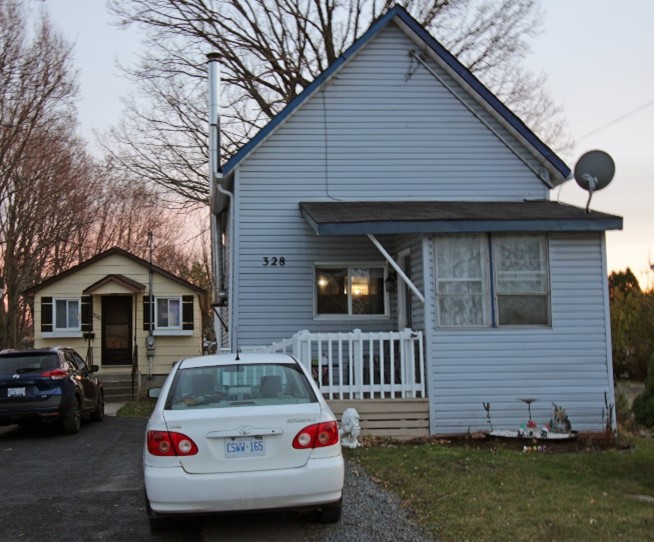 The small house at 326 5th St W was built as a bakery next to the owner’s home at 328. Small gray house with a front porch and white railing, a white car parked in the driveway. A smaller cream outbuilding is visible in the background.